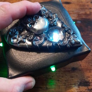 May include: A black, triangular-shaped decorative object with a silver skull and orb design. The orb glows with a blue light. The object is held in a hand.