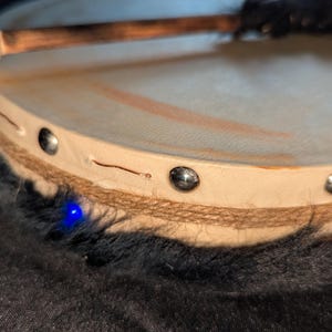 May include: A small, round drum with a light tan drumhead and a wooden mallet. The drum's rim is adorned with metal studs and wrapped with a natural fiber. A blue light is visible on the side of the drum.