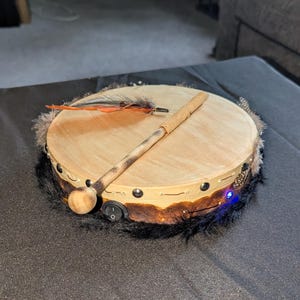 May include: A round, light-brown drum with a wooden beater and decorative feathers. The drum has a dark fur trim and a small switch. The beater is resting on the drum's surface.