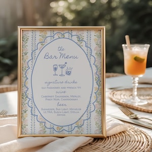 May include: A framed bar menu with a gold border, featuring a floral and blue design. The menu lists signature drinks, wine, and beer options. A cocktail glass with a straw and a slice of lime is visible in the background.
