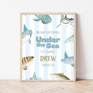 Op de afbeelding: Een ingelijste verjaardagsfeestbord met een blauw-wit gestreepte achtergrond. Het bord bevat aquarel illustraties van haaien en de tekst "Welcome to My Birthday Under the Sea Let's Celebrate Drew Turning Five."