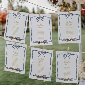 May include: Six white cards with blue borders and gold text are clipped to a string. Each card has a bow at the top and a floral design at the bottom. The cards are labeled "Table One" through "Table Six".