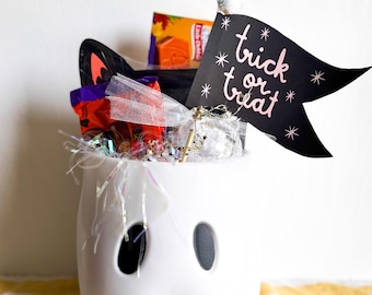 May include: A white ceramic ghost-shaped container filled with Halloween treats. The container has two oval eye cutouts and a mouth-like shape at the bottom. A black flag with "trick or treat" in pink script is attached to the top.