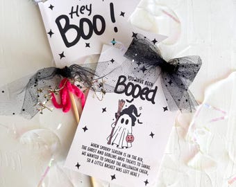 May include: Halloween-themed paper flags with the phrases "Hey Boo!" and "You have been Booed." The flags feature star accents and a ghost illustration with text about sharing treats. Black tulle bows and pink accents complete the festive decorations.