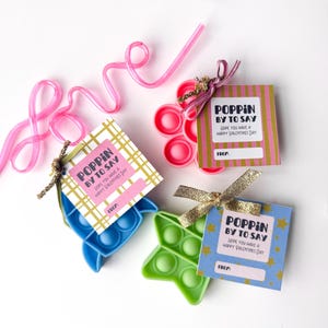 May include: Valentine's Day gifts featuring colorful pop-it toys in blue, green, and pink, each with a tag that reads "Poppin' by to say, hope you have a happy Valentine's Day." A pink straw spells out "love".