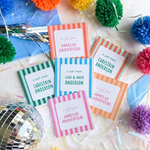 May include: A collection of square gift tags with colorful vertical stripes and white centers featuring the text a gift from followed by names like Amelia Anderson, Christain Anderson, and Luke & Owen Anderson, surrounded by yarn pom-poms and a disco ball.