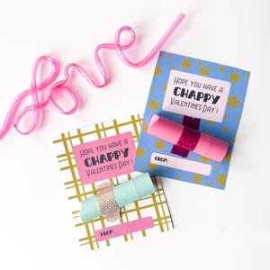May include: Two Valentine's Day cards with lip balm. One card is blue with gold stars and the text "Hope you have a Chappy Valentine's Day!". The other card is white with a gold grid pattern and the same text. A pink straw in the shape of the word "love" is also present.