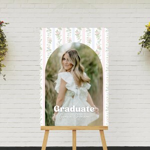 Op de afbeelding: Een afstudeerbord met een vrouw in een witte jurk, met de tekst "Graduate Gracie Carriere". Het bord heeft een roze, witte en blauwe gestreepte achtergrond met bloemaccenten en wordt weergegeven op een houten ezel.