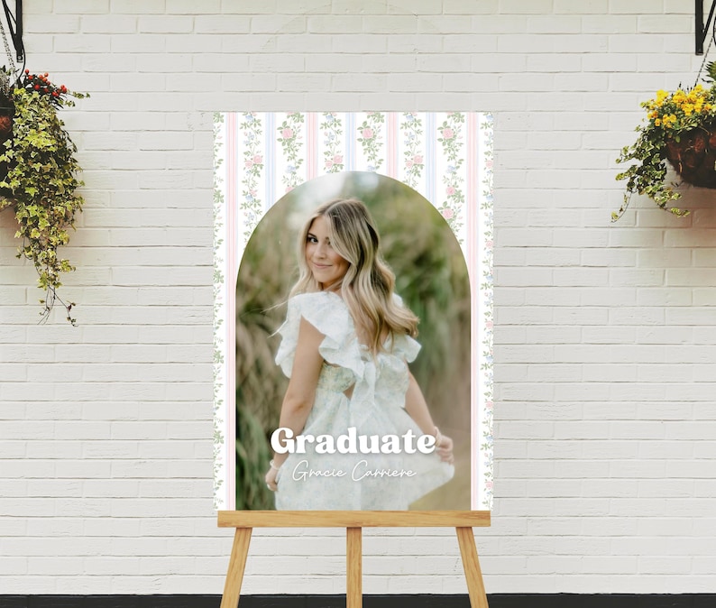 Op de afbeelding: Een afstudeerbord met een vrouw in een witte jurk, met de tekst "Graduate Gracie Carriere". Het bord heeft een roze, witte en blauwe gestreepte achtergrond met bloemaccenten en wordt weergegeven op een houten ezel.