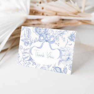 May include: A blue and white floral "Thank You" card. The card features a floral design with a bow and the words "Thank You" in a script font. The card is on a white surface with dried flowers in the background.