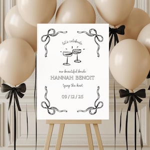 May include: A white sign with black text and illustrations announcing a wedding. The text reads "Let's celebrate our beautiful bride Hannah Benoit tying the knot 09/12/25". The sign is on an easel and surrounded by beige balloons with black ribbons.