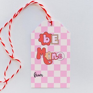 May include: Pink and white checkered gift tag with the text "Be Mine" in red and pink letters. The tag has a red and white twine string attached to it. The tag also has the word "from" written in black at the bottom.