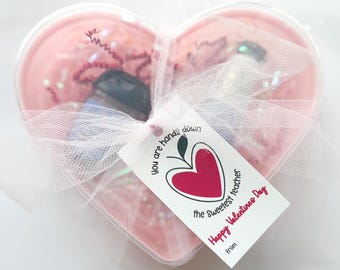 Puede incluir: Un recipiente en forma de corazón lleno de confeti rosa y objetos pequeños, envuelto en tul blanco. Una etiqueta con una manzana roja en forma de corazón y el texto "you are hands down the sweetest teacher Happy Valentines Day" está adjunta.