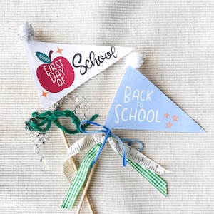 Op de afbeelding: Twee driehoekige schoolvlaggetjes. Eén witte vlaggetje leest "Happy First Day of School" met een rode appel. De andere is lichtblauw en zegt "Back to School". Beide hebben pompons en zijn met linten aan houten stokjes bevestigd.