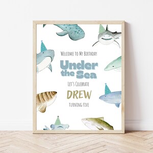 Op de afbeelding: Een ingelijste print met een lichte houten afwerking. De print toont aquarel illustraties van haaien met feesthoeden, met de tekst "Welcome to My Birthday, Under the Sea, Let's Celebrate DREW Turning Five."