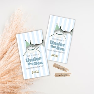 Puede incluir: Dos tarjetas rectangulares con un fondo a rayas azul claro y blanco. Cada tarjeta presenta un tiburón de dibujos animados con un gorro de fiesta. El texto en las tarjetas dice "Gracias por unirte a mí Bajo el mar, ¡Hiciste que mi cumpleaños fuera genial!" y "Drew".