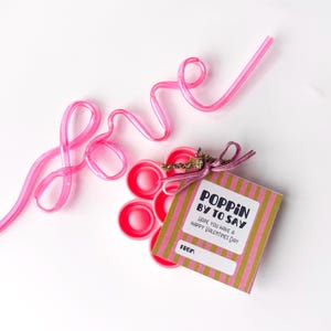 May include: Pink plastic straw shaped into the word "love" next to a stack of pink bottle caps and a Valentine's Day gift tag. The tag reads "Poppin' by to say, Hope you have a Happy Valentine's Day."