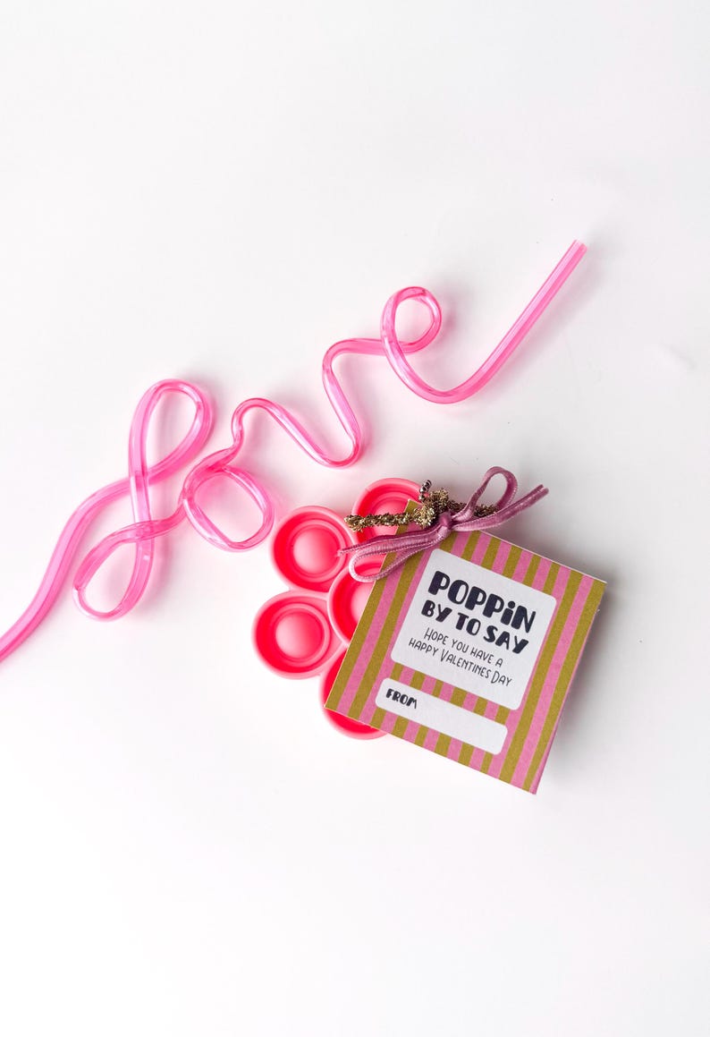 May include: Pink plastic straw shaped into the word "love" next to a stack of pink bottle caps and a Valentine's Day gift tag. The tag reads "Poppin' by to say, Hope you have a Happy Valentine's Day."