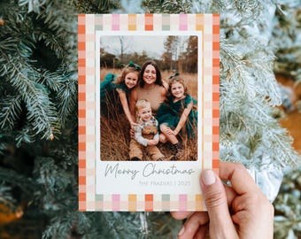 May include: A Christmas card with a family photo inside a white border, set against a colorful plaid pattern. The card reads "Merry Christmas" with "THE FRAZIERS | 2025" below. The card is held in a hand.