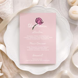 Może przedstawiać: Różowa karta menu z kwiatową ilustracją i napisem "the Menu". Menu zawiera przystawkę, danie główne i deser, ze szczegółowymi opisami. Karta leży na białym talerzu z falistymi krawędziami i elementami dekoracyjnymi.