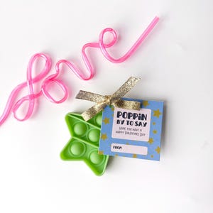 May include: A green star-shaped pop-it toy with a blue tag that says "Poppin' by to say, Hope you have a Happy Valentines Day". A pink straw spells out the word "love". A gold ribbon is tied to the tag.