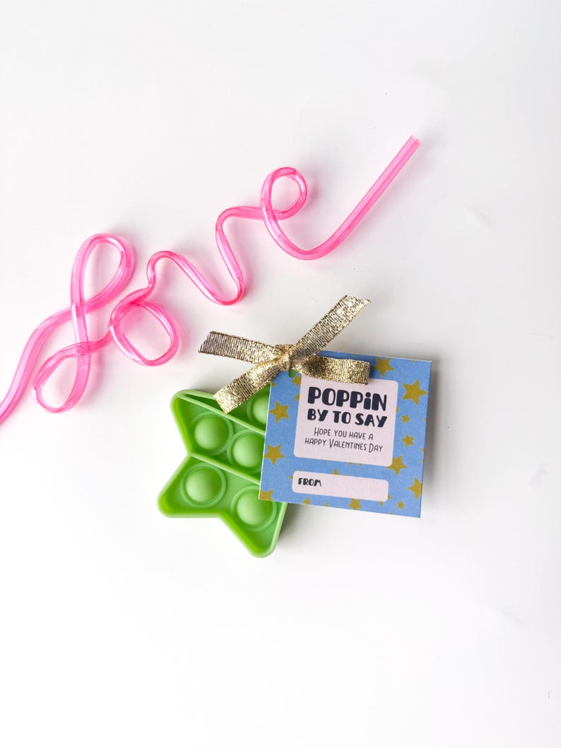 May include: A green star-shaped pop-it toy with a blue tag that says "Poppin' by to say, Hope you have a Happy Valentines Day". A pink straw spells out the word "love". A gold ribbon is tied to the tag.