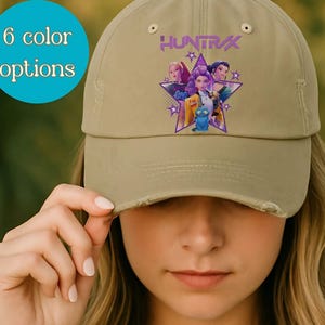 May include: A khaki baseball cap featuring a purple star design with the word "HUNTRAX" and images of characters. The cap has a distressed brim and the text "6 color options" in a blue circle.