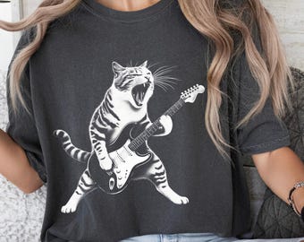 May include: A dark grey t-shirt featuring a white graphic of a cat playing an electric guitar. The cat is singing with its mouth open and has a striped tail.