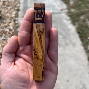 May include: A wooden mezuzah with a dark brown wood finish and a carved Hebrew symbol. The mezuzah is about 6 inches long and 1 inch wide.