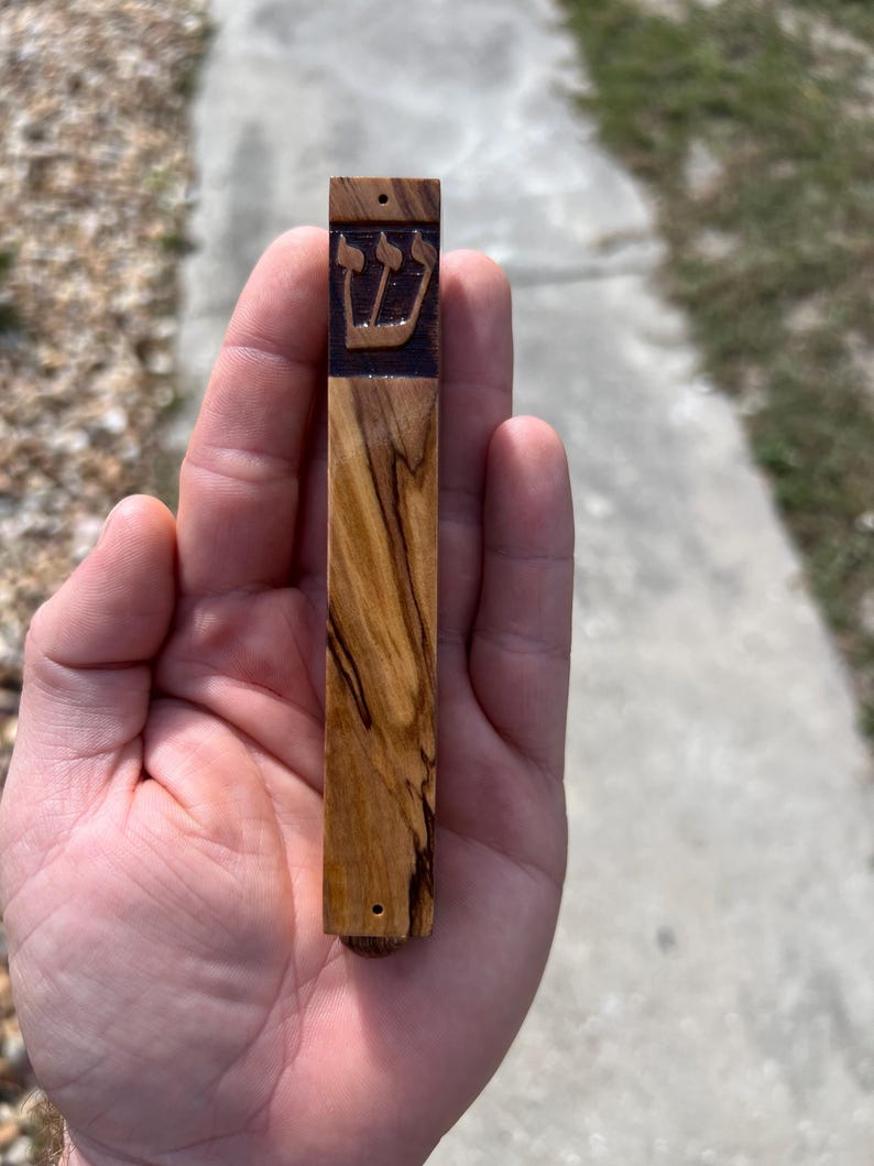 May include: A wooden mezuzah with a dark brown wood finish and a carved Hebrew symbol. The mezuzah is about 6 inches long and 1 inch wide.