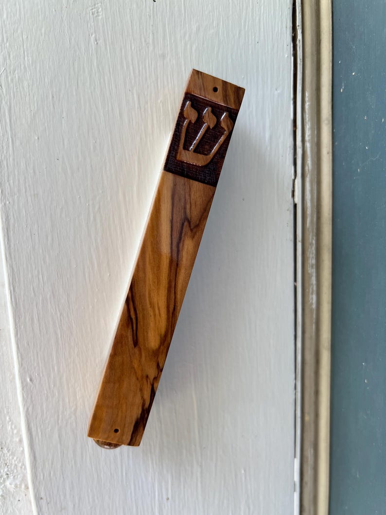 May include: A wooden mezuzah with a carved Star of David design. The mezuzah is attached to a white door frame.