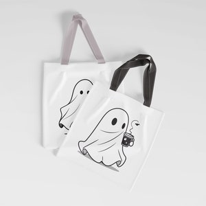 May include: Two white tote bags with black handles. One bag features a ghost holding a cup of coffee with a bat flying above. The other bag features a ghost with a simple design.