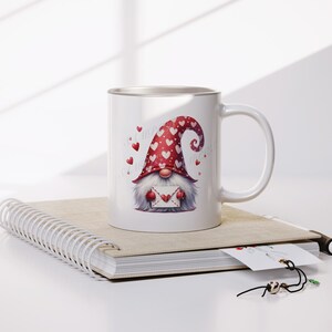 May include: White ceramic mug with a red and white gnome holding a heart-shaped envelope. The gnome is wearing a red hat with white hearts. The mug is sitting on a spiral bound notebook.