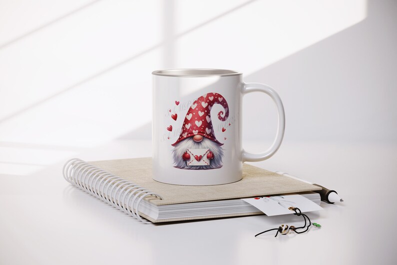 May include: White ceramic mug with a red and white gnome holding a heart-shaped envelope. The gnome is wearing a red hat with white hearts. The mug is sitting on a spiral bound notebook.