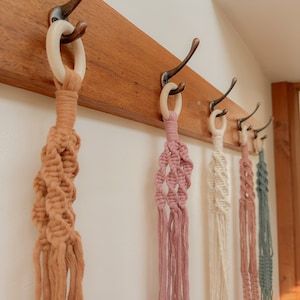 May include: Four macrame plant hangers in different colours hanging on a wooden rack with hooks. The hangers are made of natural fibres and have a simple, bohemian style.