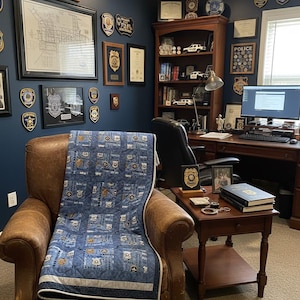 May include: A cozy home office with a brown leather armchair draped with a blue quilt featuring police-themed patches. The room is decorated with framed badges, a map, and an American flag. A wooden desk and bookshelf complete the space.