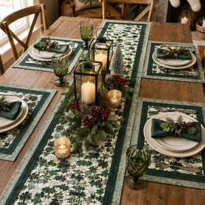 May include: A dining table set featuring a long table runner with a green and white botanical pattern, matching placemats, dark green cloth napkins with pinecone and berry accents, white plates, green glassware, and black metal lanterns.