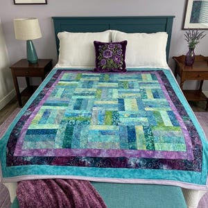 May include: A handmade quilt with a patchwork design in shades of blue, green, and purple. The quilt is on a bed with white pillows and a decorative purple pillow. A matching throw blanket is draped over a bench at the foot of the bed.