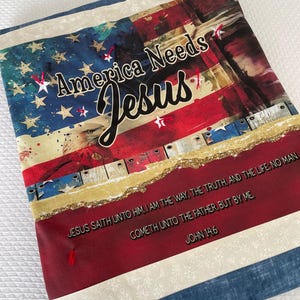 May include: A rectangular fabric item featuring a design incorporating the American flag, with the words "America Needs Jesus" and a Bible verse. The design includes red, white, and blue colors, with a distressed aesthetic.