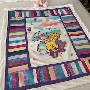 May include: A white and purple quilted baby blanket with a Winnie the Pooh and friends design. The design features Winnie the Pooh, Tigger, Piglet, Eeyore, and a blue character holding an umbrella. The blanket has a white border and a purple and white striped border.