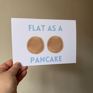 May include: A white card with blue text that reads "FLAT AS A" and two brown pancake illustrations. The text "PANCAKE" is also in blue below the pancakes.