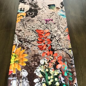 May include: A colorful table runner with a floral and butterfly design. The runner features a variety of flowers, butterflies, and other natural elements in shades of red, orange, yellow, green, blue, and purple. The background is a light brown color.