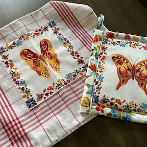 Puede incluir: Un paño de cocina a cuadros blancos y rojos y un salvamanteles a juego. Ambos presentan un diseño cuadrado con una ilustración de mariposa en tonos naranja, amarillo y rojo, rodeada por un borde floral.