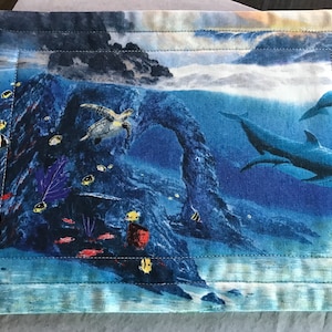 Può includere: Un tovaglietta all'americana trapuntata blu e bianca con una scena sottomarina colorata che presenta una tartaruga marina, delfini e pesci. La tovaglietta ha un bordo cucito.