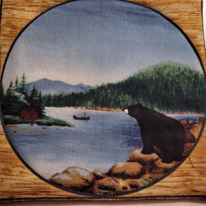 Puede incluir: Un posavasos cuadrado de tela con una imagen circular de una escena de lago. Un oso negro está sentado en rocas, con vista a un lago con una canoa y una pequeña cabaña. El borde tiene un patrón de vetas de madera.