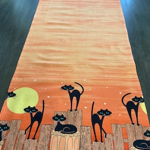 May include: A rectangular table runner featuring an orange gradient background with a pattern of black cats perched on wooden fences and stone pillars. The design includes a large yellow moon and small white stars scattered across the fabric.