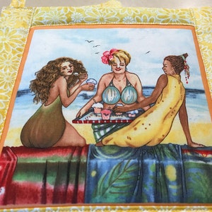 Op de afbeelding: Drie vrouwen zitten aan een tafel op een strand en eten cupcakes. De vrouw in het midden draagt een blauw-groen badpak en een roze bloem in haar haar. De vrouw links draagt een groen badpak en de vrouw rechts draagt een gele jurk. De tafel is bedekt met een rood-wit geblokt tafelkleed.