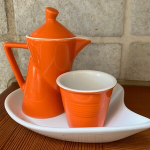 May include: A bright orange ceramic coffee set with a matching creamer, cup, and serving tray. The set includes a small, cylindrical cup and a creamer with a lid. The serving tray is white and has a unique, curved shape.