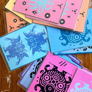 Puede incluir: Una colección de coloridas impresiones artísticas con varios diseños de monstruos. Las impresiones son en tonos de rosa, azul, naranja y amarillo, con ilustraciones en negro y verde azulado. Las impresiones son rectangulares y parecen estar empaquetadas en plástico transparente.