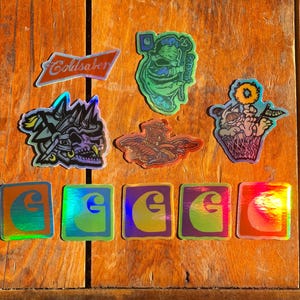 May include: A selection of holographic stickers on a wooden surface. The stickers display diverse designs, including a skull, an angel, a cartoon character in a waffle cone, and the Carhartt logo. One sticker reads "Coldsaber".